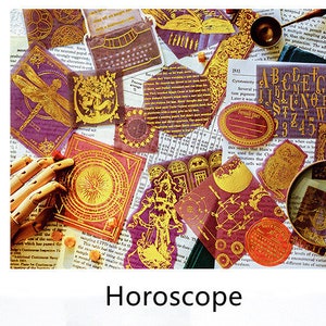 May include: A collection of decorative stickers and ephemera with a horoscope theme. The stickers feature gold foil accents on a burgundy background, with images of a dragonfly, sun, and other celestial designs. A vintage typewriter is also present.