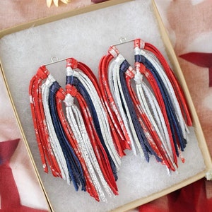 May include: A pair of red, white, and blue leather tassel earrings, presented in a small box. The earrings feature a silver-tone metal finding and are arranged in a patriotic color scheme.