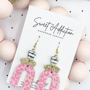 May include: Pink and gold statement earrings with a leopard print design and a white and black striped accent. The earrings are handmade and feature a gold hook closure. The earrings are displayed on a white card with the text "Sweet Addiction Handmade Jewelry".