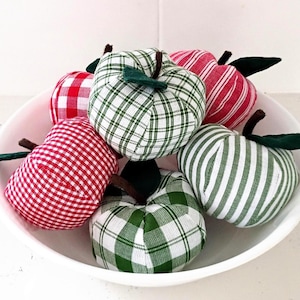May include: A collection of fabric apples in a white bowl, featuring various patterns including red and white gingham, green and white plaid, red and white stripes, and green and white stripes. Each apple has a dark green fabric leaf and a brown stem.