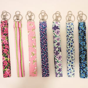 以下が含まれることがあります： ピンク、紫、青などの色合いで、それぞれ異なる花柄やストライプ柄が施された7本の布製リストレットキーホルダーが並んでいます。各ストラップには、銀色の金属製ロブスタークラスプと円形の二重リングが取り付けられています。