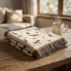 May include: A cream-colored woven throw blanket featuring a repeating pattern of dark brown hunting dogs and game birds. The blanket has a textured fabric construction and is finished with multi-colored fringed edges on the short sides.