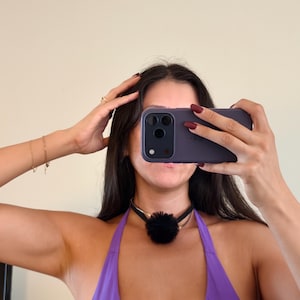 May include: A dark purple smartphone held in a hand, featuring a triple-lens camera system. A black choker necklace with a prominent, round, fluffy black pom-pom charm is worn around the neck, paired with a bright purple halter-style garment.