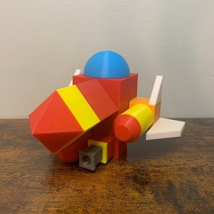 May include: A red, yellow, and white spaceship toy made of blocks. The spaceship has a blue dome on top and a white wing on the side.