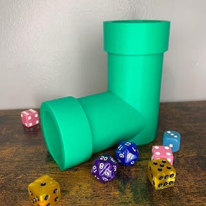 May include: A green plastic pipe-shaped dice tower with six dice of various colors and numbers on a wooden surface.