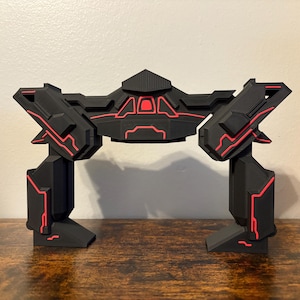 May include: A black and red geometric sculpture with a futuristic design. The sculpture has a central structure with two upright supports. Red lines accent the edges and interior of the black structure. The sculpture is on a wooden surface.