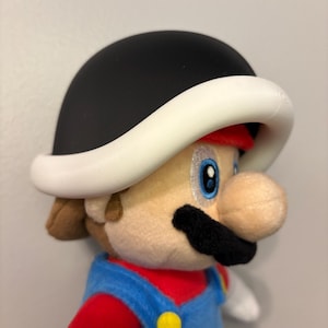 May include: A plush toy of the video game character Mario wearing a black and white Koopa shell helmet. Mario is wearing his signature red shirt, blue overalls, and has a black mustache.