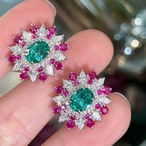 May include: A pair of gemstone earrings featuring a central oval-cut emerald surrounded by a halo of pink and white gemstones. The earrings have a floral design, with the pink gemstones forming the outer petals and the white gemstones creating a starburst effect.