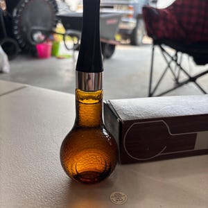 May include: An amber glass bottle with a textured surface and a long, slender black neck topped with a silver band. A rectangular box sits beside the bottle, and a small circular label on the surface reads 2 FL OZ (56 ML).