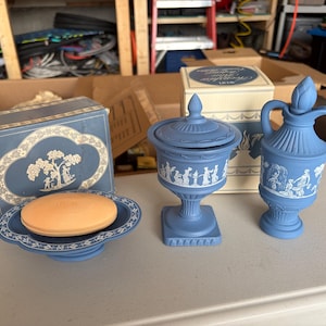 May include: A blue and white ceramic soap dish with a peach-colored soap bar, a lidded urn, and a small pitcher. Each piece features white relief figures on a matte blue background. The items are displayed with their original cardboard boxes.