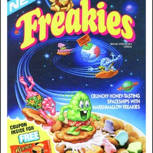 May include: A box of Ralston Freakies cereal featuring a space-themed design with cartoon characters. The box has a coupon for a free package of Reese's Pieces. The cereal is described as "Crunchy Honey-Tasting Spaceships with Marshmallow Freakies" and is fortified with 9 essential vitamins and minerals. The box is 11 ounces (311g).