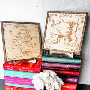 May include: Two wooden map art pieces, one labeled Prythian and the other Erilea, are displayed with a stack of books by Sarah J. Maas. The books have colorful covers and titles like "A Court of Thorns and Roses" and "Crown of Midnight."