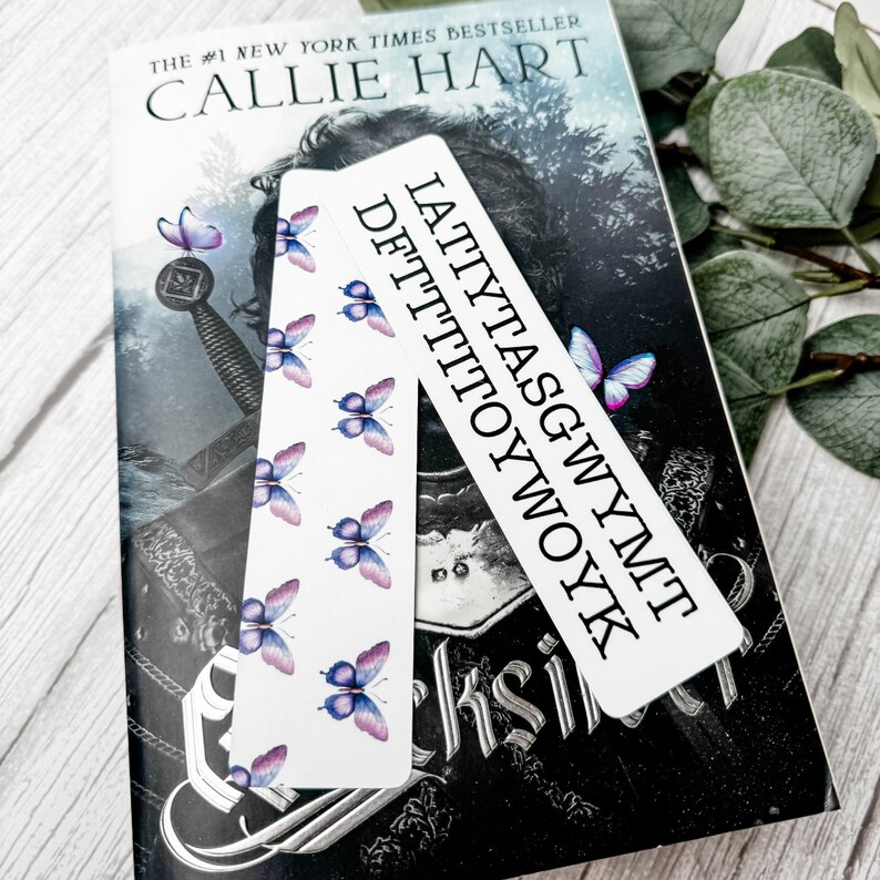 May include: Two rectangular white bookmarks featuring a repeating pattern of small purple and blue butterflies. One bookmark displays the black text IATTYTASGWYMT DFTTTTOYWOYK in a bold, sans-serif font, while the other shows only the butterfly design.
