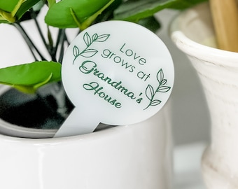 May include: A white plant marker with the text "Love grows at Grandma's House" in a green script font. The marker has a circular top with leaf designs and a rectangular stem, placed in a white ceramic pot with green leaves.