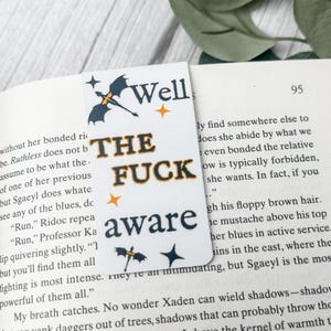 May include: White bookmark with the text "Well THE FUCK aware" in black and orange. The bookmark features a bat and dragon graphic and star accents. The bookmark is placed in a book.