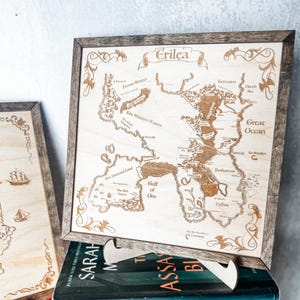 May include: Wooden map of Erilea, framed in a rustic style. The map features detailed geographical elements, including the Great Ocean, Gulf of Oro, and various regions. The text "Erilea" is displayed at the top. The map is resting on a book.