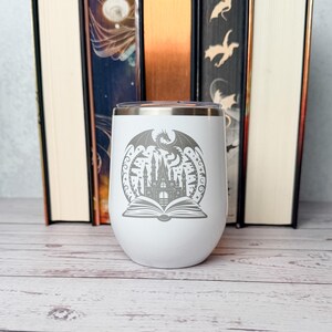 May include: A white stainless steel tumbler featuring a gray laser-engraved design of an open book with a castle rising from the pages and a dragon flying above. The tumbler has a smooth, rounded shape and a metallic rim at the top opening.
