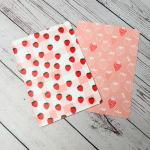 May include: Two rectangular cards with strawberry patterns. One card features red strawberries on a white and pink gingham background. The other card has a pink background with white and red strawberry outlines.