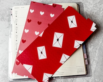 May include: Two rectangular paper bookmarks placed on an open planner. One bookmark is dark red with a white heart pattern. The other is bright red with a white envelope pattern featuring small black hearts on the envelope flaps.