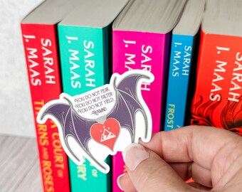 May include: A die-cut sticker featuring purple bat wings framing a red heart with a white mountain and star icon. The text reads: YOU DO NOT FEAR. YOU DO NOT FALTER. YOU DO NOT YIELD. -RHYSAND. The sticker is held against a row of colorful book spines.