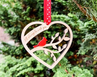 May include: Wooden heart-shaped ornament with a red cardinal perched on a branch. The ornament has the engraved text "I'll always be with you" and is suspended by a red and white polka dot ribbon. The background is a blurred green.