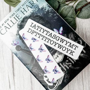 May include: A bookmark with watercolor butterflies in shades of purple and blue, and the text "IATITYASGWYMT DFTTTITOYWOYK" is placed on top of a book. The book cover features the title "Callie Hart" and is a New York Times bestseller.