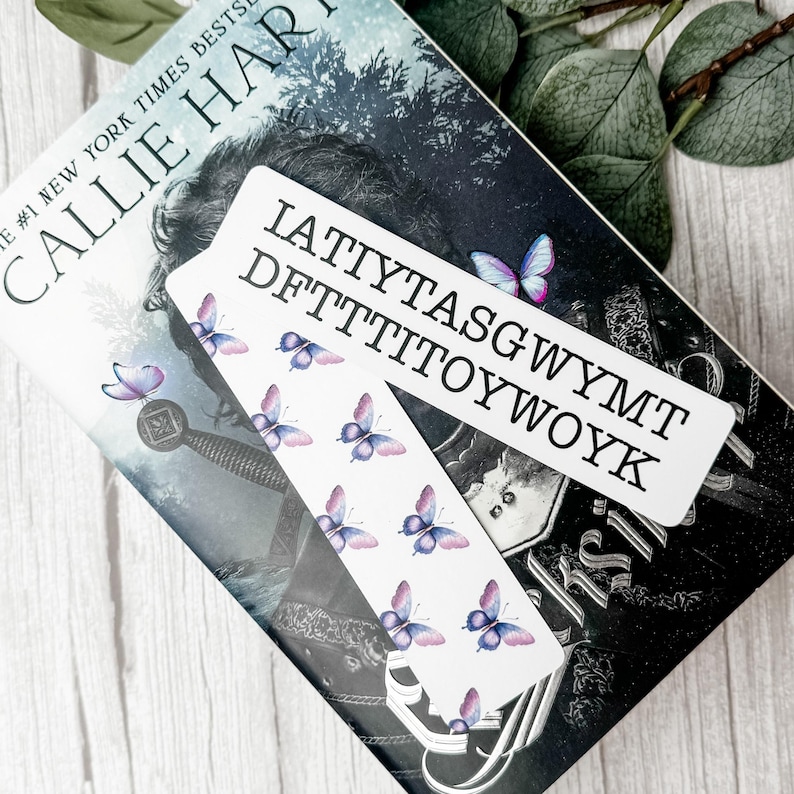May include: A rectangular white bookmark featuring a repeating pattern of small purple and blue butterflies. The text IATTYTASGWYMT DFTTTITOYWOYK is printed in black capital letters across the top section of the bookmark surface.