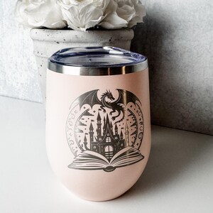 May include: A light pink stainless steel tumbler with a clear lid, featuring a dark gray laser-engraved graphic of an open book with a castle rising from the pages and a dragon flying above it, all enclosed within a circular decorative border.