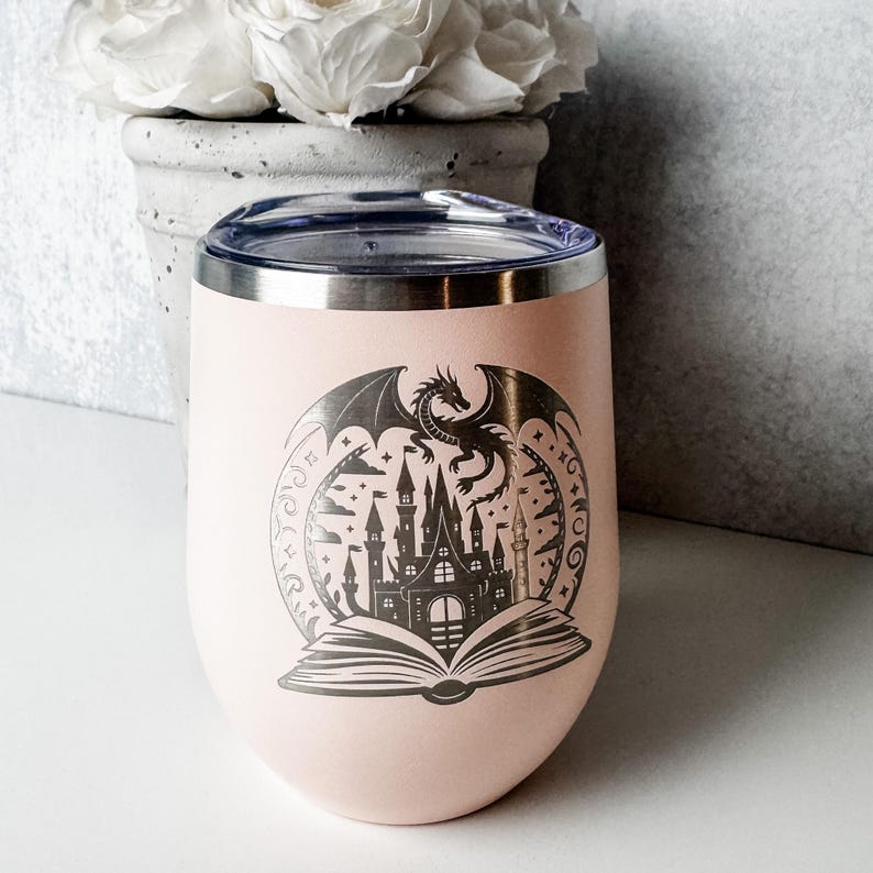 May include: A light pink stainless steel tumbler with a clear lid, featuring a dark gray laser-engraved graphic of an open book with a castle rising from the pages and a dragon flying above it, all enclosed within a circular decorative border.