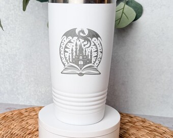 May include: A white stainless steel tumbler with a tapered cylindrical shape and horizontal ridges near the base. The front features a gray laser-engraved graphic of an open book with a castle rising from the pages and a dragon flying above.