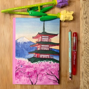Può includere: Un quaderno rosa con un'illustrazione ad acquerello di un pagoda giapponese, il monte Fuji e fiori di ciliegio. Il quaderno è su una superficie di legno con un portapenne verde e giallo e una penna rossa e verde.