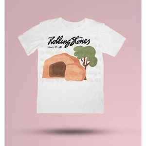 May include: White t-shirt with the Rolling Stones logo and a graphic of a tomb and tree. The text "Rolling Stones Since 33 AD" is printed above the graphic in black script.