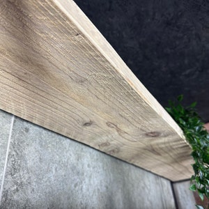 May include: Close-up of a light wood shelf with a rough, unfinished texture. The shelf is mounted to a grey tiled wall.