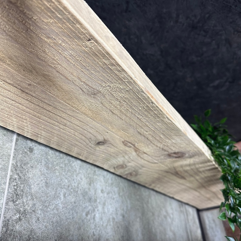 May include: Close-up of a light wood shelf with a rough, unfinished texture. The shelf is mounted to a grey tiled wall.