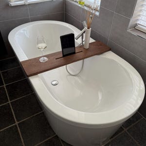 Può includere: Una vasca da bagno ovale bianca con un vassoio da bagno in legno scuro che contiene un tablet, un bicchiere di vino bianco e un vaso di ceramica bianca con canne. Il bagno ha pareti piastrellate grigie e un pavimento piastrellato scuro.
