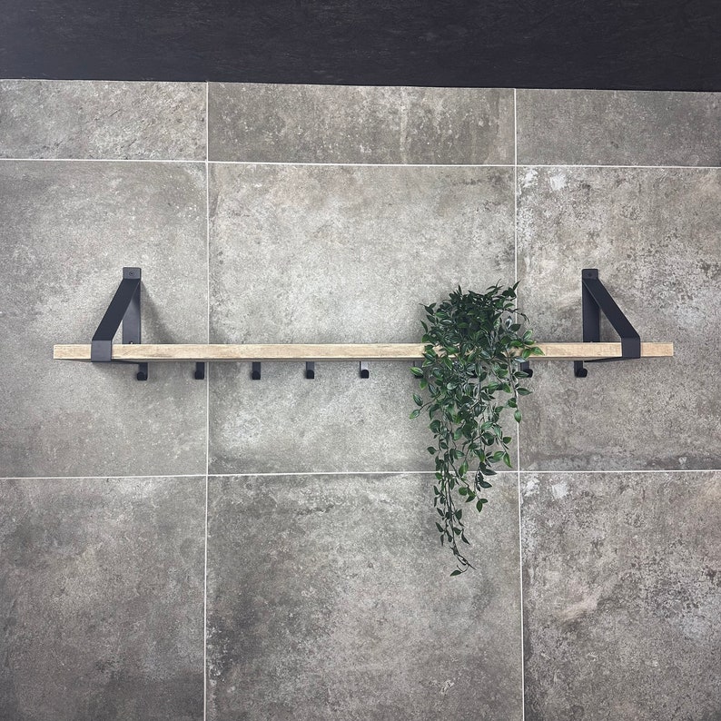 Puede incluir: Una estanter&iacute;a de madera con soportes y ganchos de metal negro, montada en una pared de azulejos grises. Una planta verde cuelga de la estanter&iacute;a.