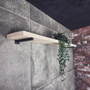 Puede incluir: Una estantería de madera con soportes de metal negro montados en una pared de azulejos grises. Una planta en maceta con hojas verdes colgantes cuelga de la estantería.