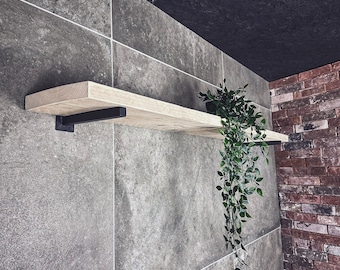 May include: A wooden shelf with black metal brackets mounted on a grey tiled wall. A potted plant with trailing green leaves hangs from the shelf.