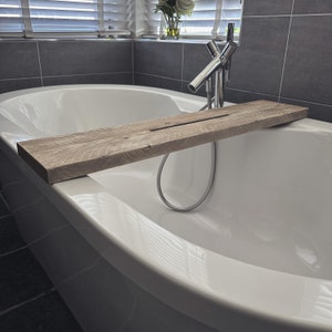 Puede incluir: Una ba&ntilde;era blanca con una bandeja de ba&ntilde;o de madera. La bandeja est&aacute; hecha de madera natural y tiene una ranura para sujetar una botella u otros art&iacute;culos. La ba&ntilde;era est&aacute; en un ba&ntilde;o moderno con paredes de azulejos grises.