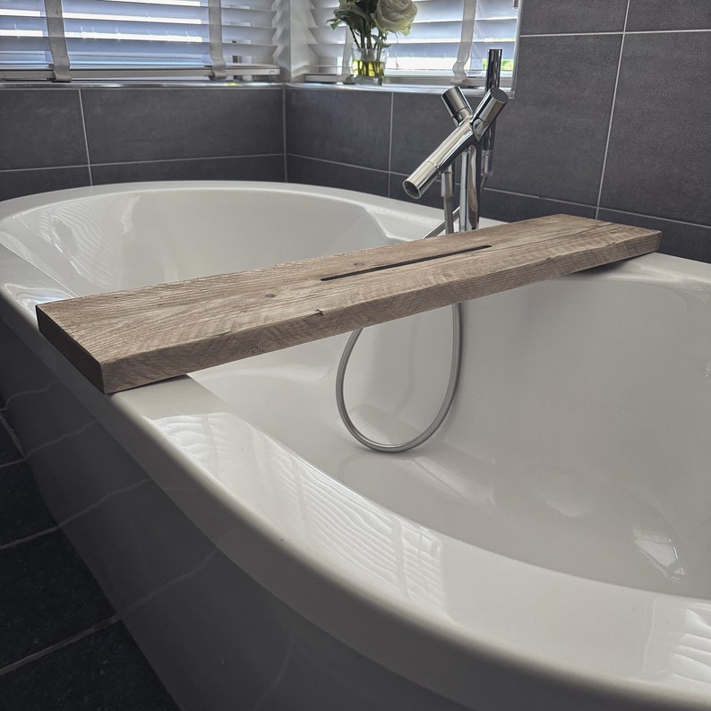 Puede incluir: Una ba&ntilde;era blanca con una bandeja de ba&ntilde;o de madera. La bandeja est&aacute; hecha de madera natural y tiene una ranura para sujetar una botella u otros art&iacute;culos. La ba&ntilde;era est&aacute; en un ba&ntilde;o moderno con paredes de azulejos grises.