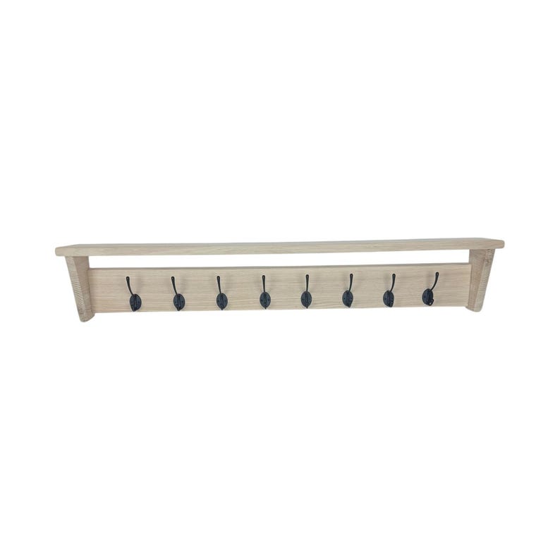 Puede incluir: Una estanter&iacute;a de pared de madera de color claro con ocho ganchos de metal negro. La estanter&iacute;a tiene un dise&ntilde;o rectangular simple con una balda superior para guardar cosas. Los ganchos est&aacute;n espaciados uniformemente a lo largo del borde inferior, proporcionando una soluci&oacute;n de almacenamiento funcional y decorativa.