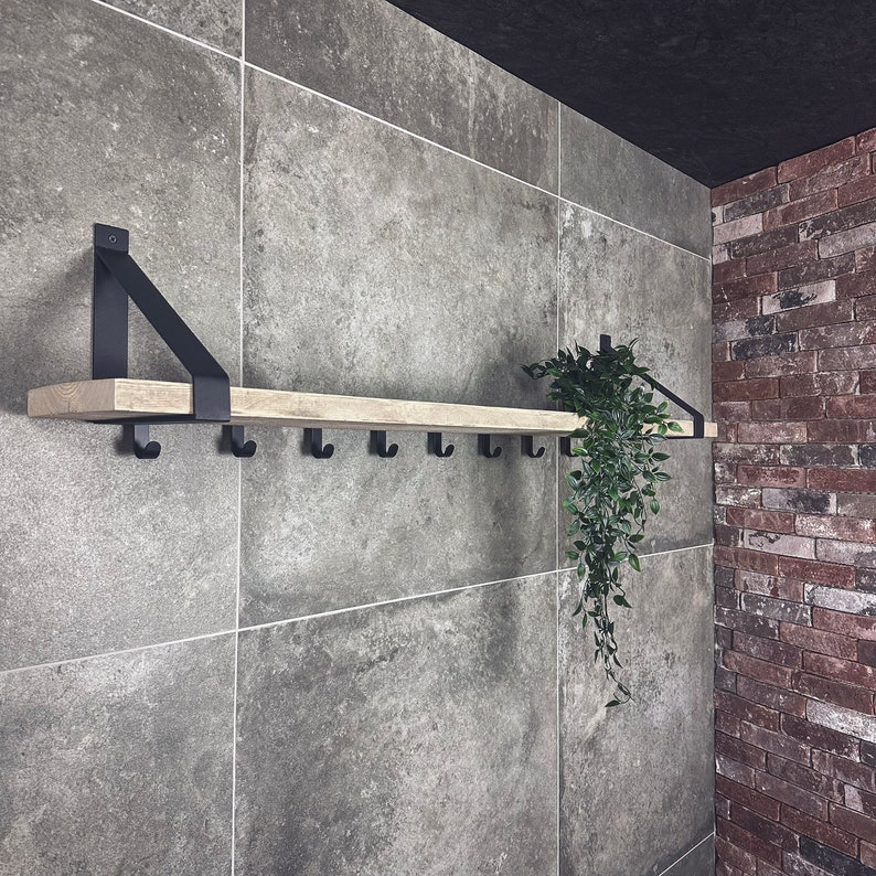 Puede incluir: Una estanter&iacute;a de madera con soportes y ganchos de metal negro, montada en una pared de azulejos grises. Una planta verde cuelga de la estanter&iacute;a.