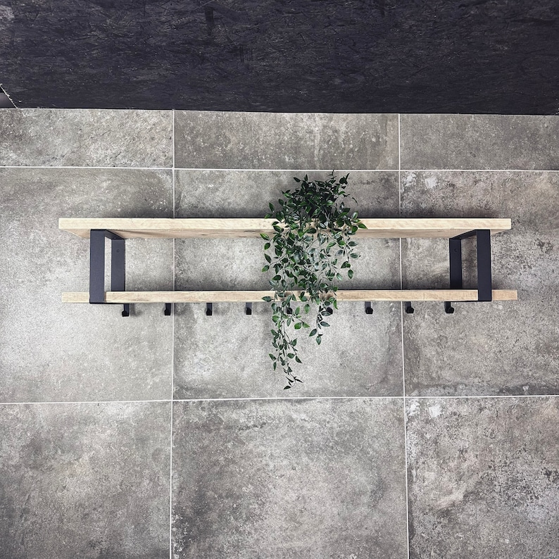 May include: A two-tiered wooden shelf with black metal brackets and hooks. The shelf is mounted on a grey tiled wall. A green plant hangs in front of the shelf.