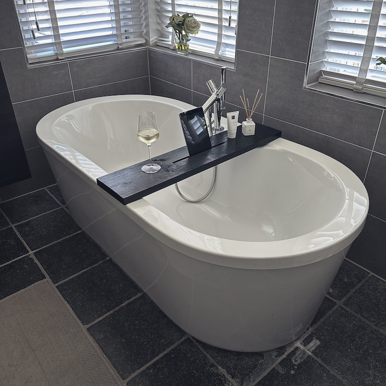 May include: A white bathtub with a wooden tray and a glass of wine. The tray has a tablet, a bottle of lotion, and a bottle of essential oil.