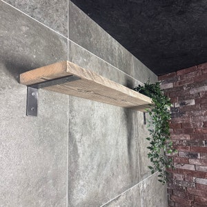Puede incluir: Una estantería de madera rústica con soportes metálicos, montada en una pared de azulejos grises. Una planta verde cuelga de la estantería.