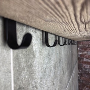 May include: A row of black metal hooks mounted under a wooden shelf. The hooks are attached to a grey tiled wall.