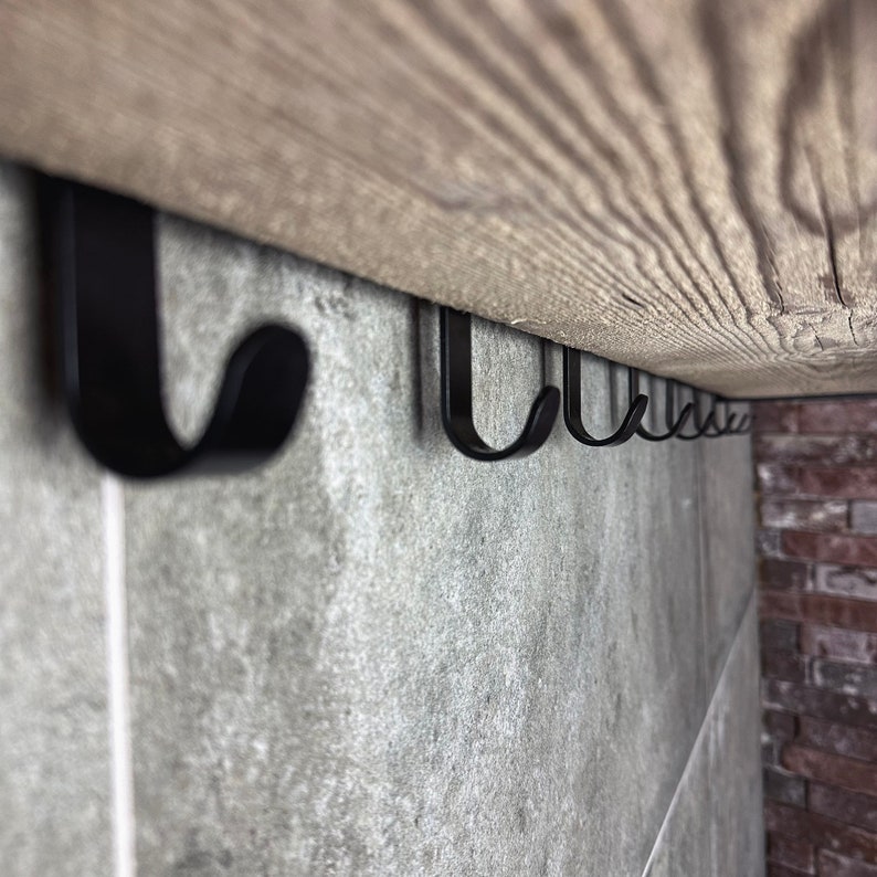 May include: A row of black metal hooks mounted under a wooden shelf. The hooks are attached to a grey tiled wall.