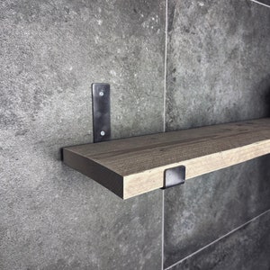 Puede incluir: Una estantería de madera con soportes de metal negro. La estantería está montada en una pared de azulejos grises.