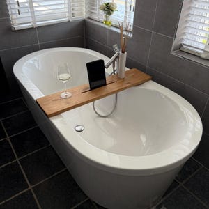 Könnte beinhalten: Eine weiße, ovale Badewanne mit einem hölzernen Badewannen-Tablett, auf dem sich ein Tablet, ein Weinglas und eine kleine Vase befinden. Das Badezimmer hat graue Fliesenwände und ein Fenster mit weißen Jalousien. Thema: Entspannung.