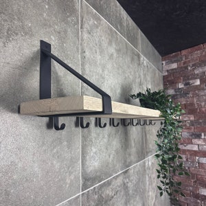 Puede incluir: Una estanter&iacute;a de madera con soportes y ganchos de metal negro. La estanter&iacute;a est&aacute; montada en una pared de azulejos grises. Una peque&ntilde;a planta en maceta cuelga de la estanter&iacute;a.